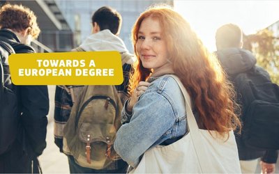 Eine Studentin, die fröhlich in die Kamera blickt und im Hintergrund drei Studierende, die einen Weg entlanggehen. 
